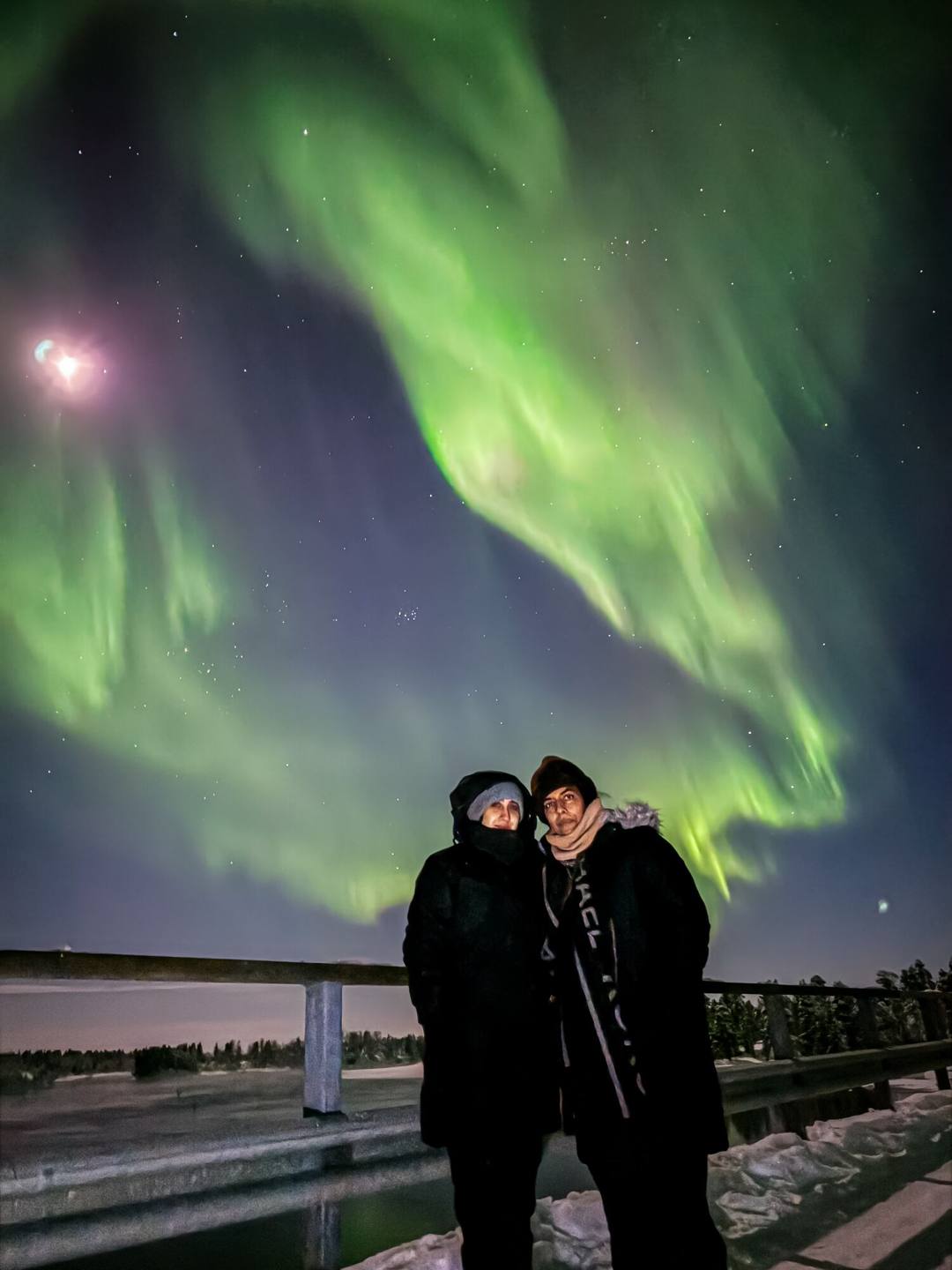 Two guests on a bridge under green Northern Lights and a bright moon