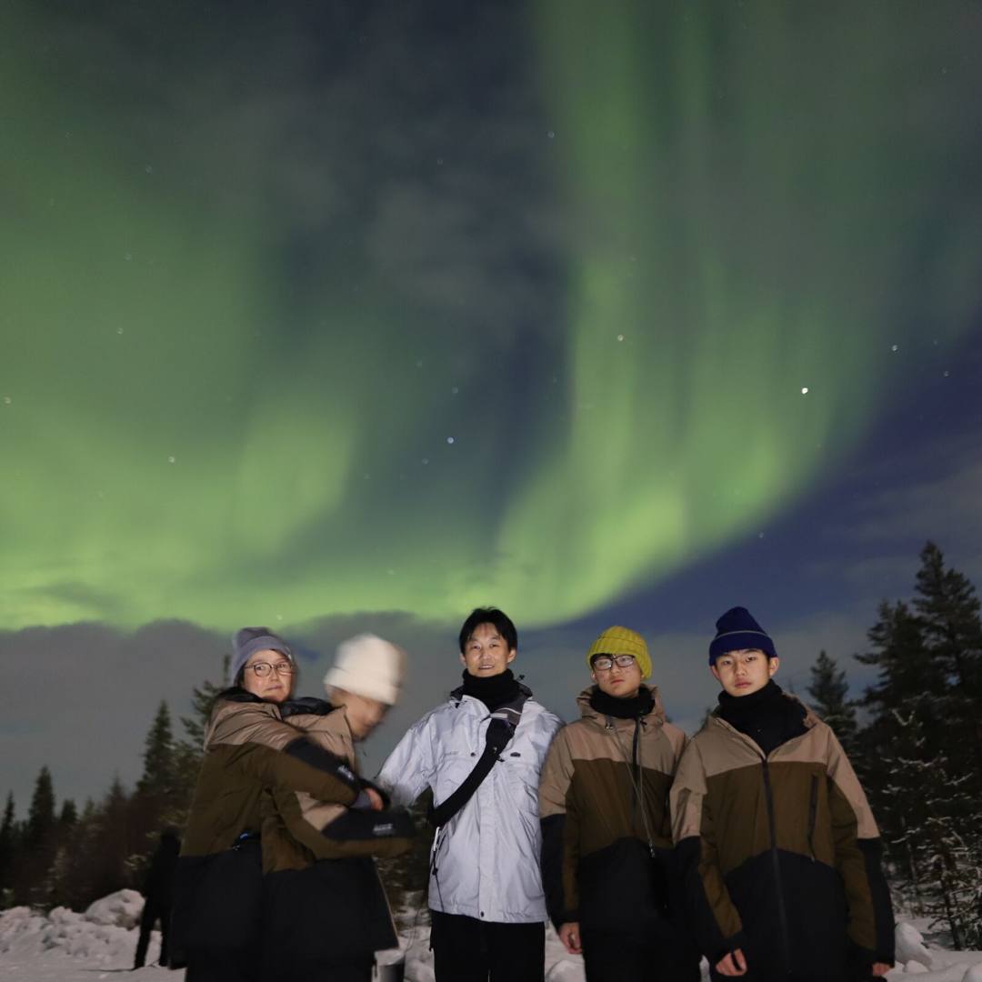 Group of five guests under green Northern Lights by snowy trees