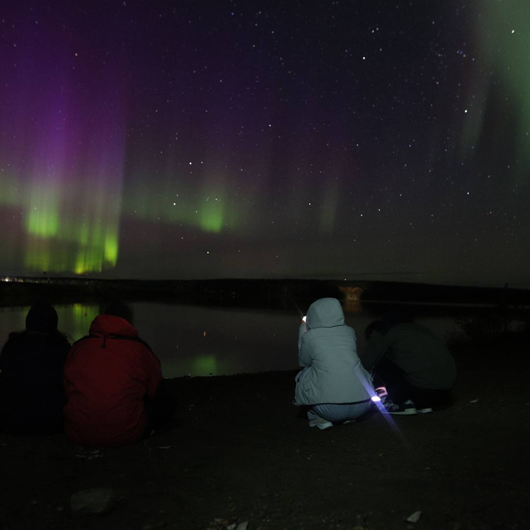 Guests sitting by a lake watching green and purple Northern Lights