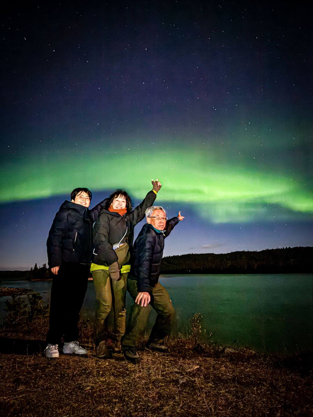 Three guests pointing at a green aurora arc on a lakeshore