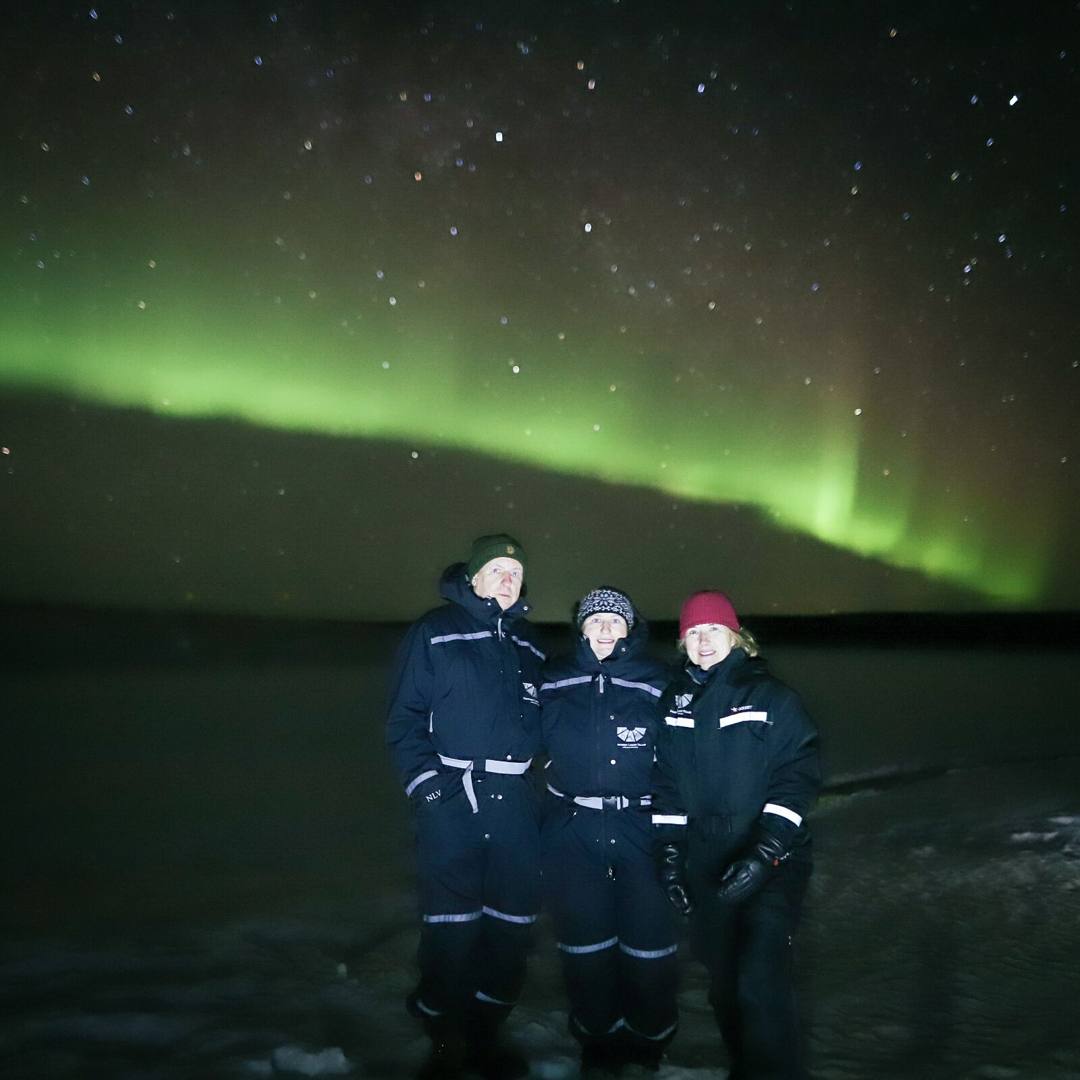Three guests in thermal overalls under a green aurora arc