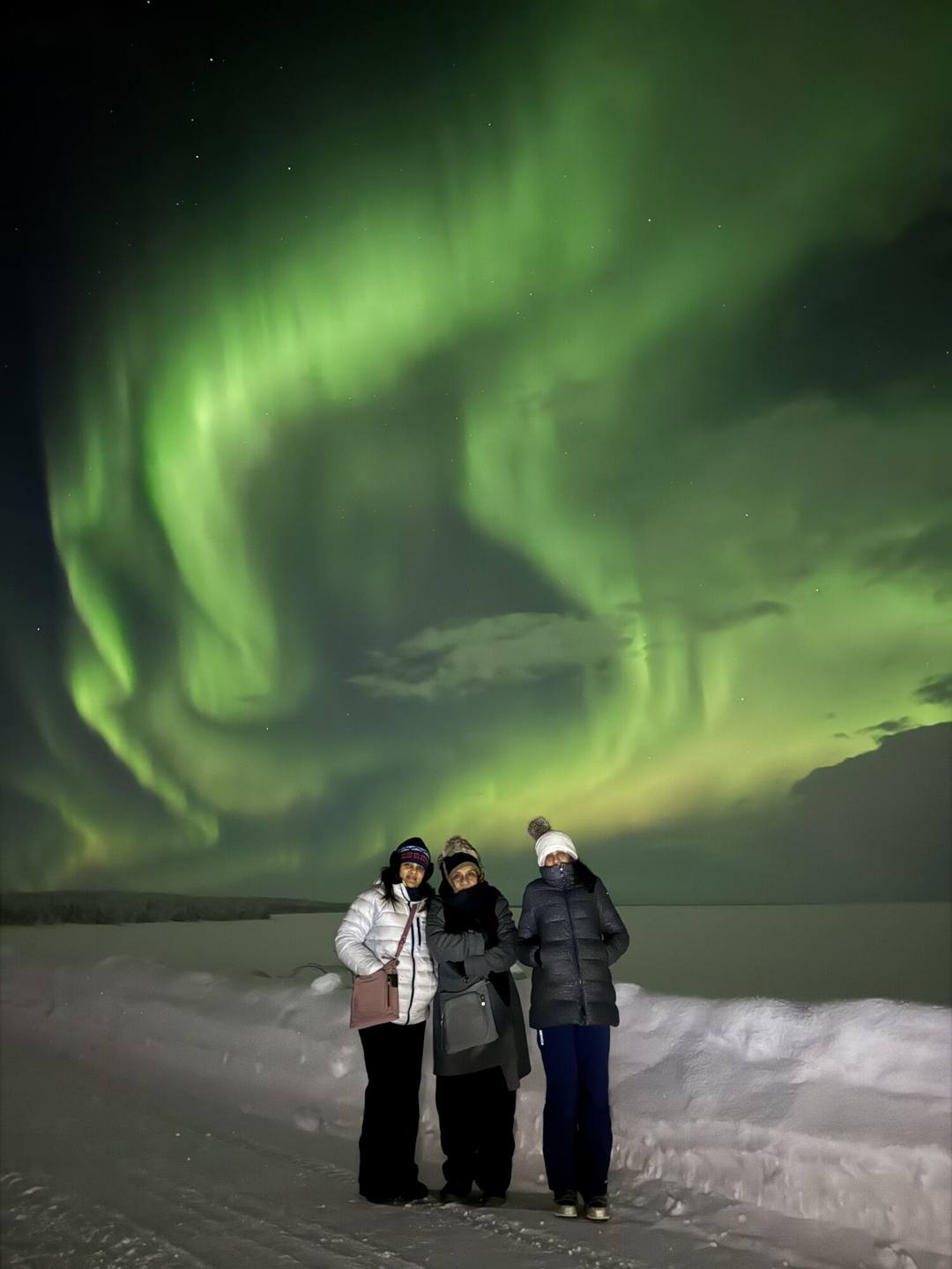 Three guests on a snowy lake under swirling green Northern Lights
