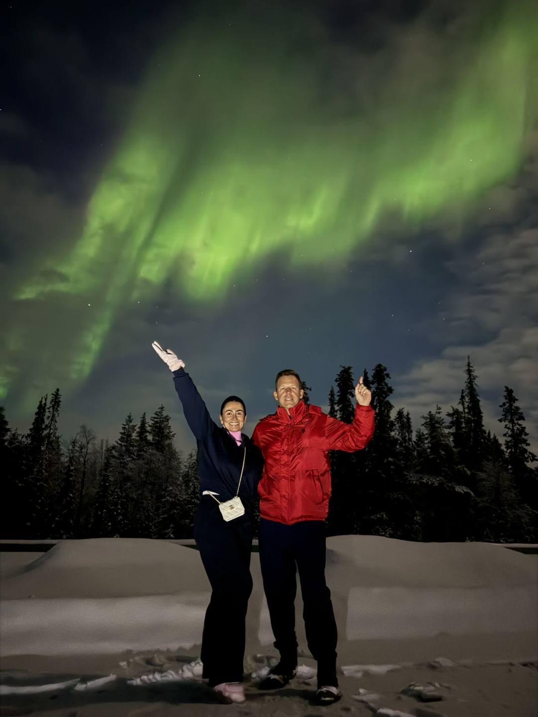 Two guests waving under green Northern Lights beside snowy forest