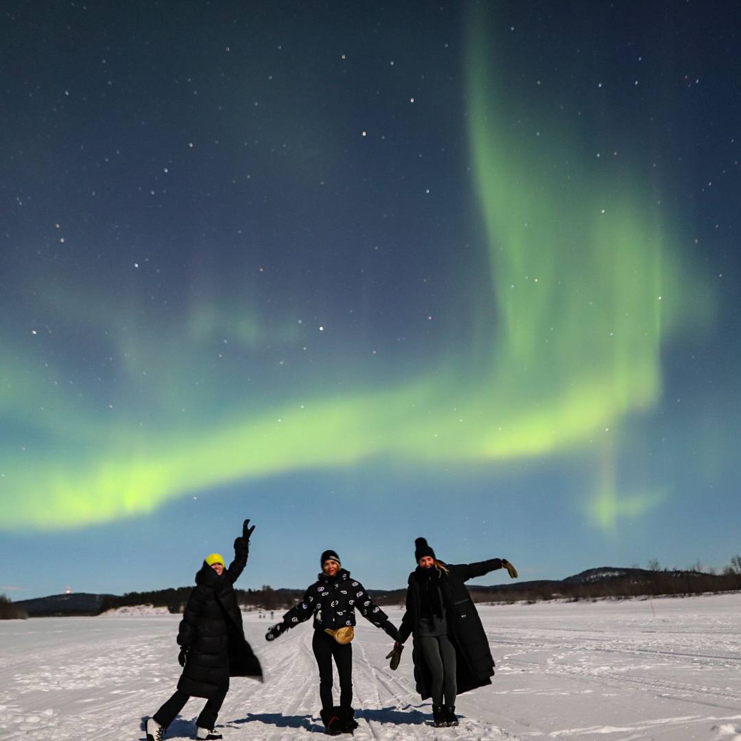 Three people posing on a snowy frozen lake under Northern Lights