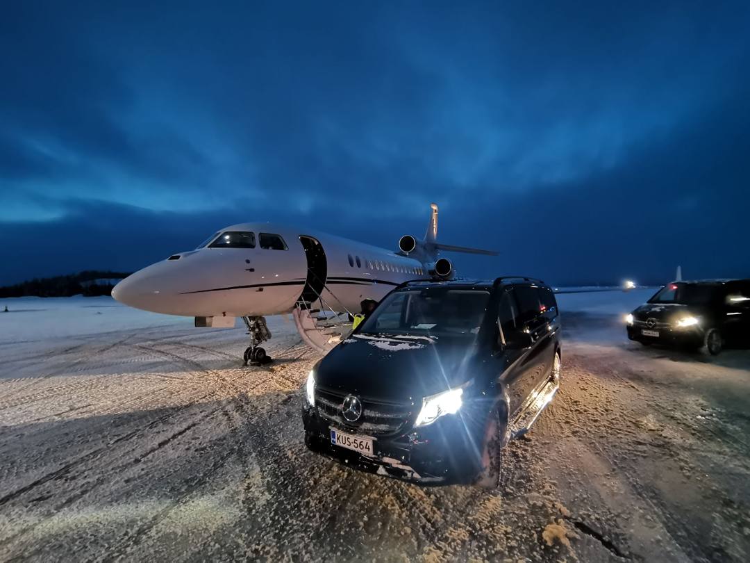 Black Mercedes-Benz van with headlights on beside a private jet on a snowy airport apron at dusk