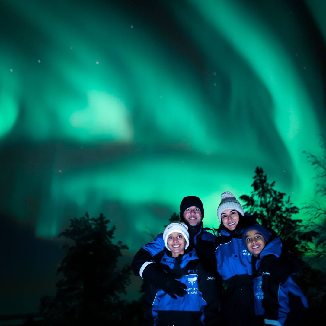 Group of four guests in winter overalls smiling under green Northern Lights